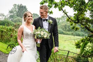 Newlyweds share a joyful moment walking together on a rainy day in the countryside, with a beautiful white bouquet.