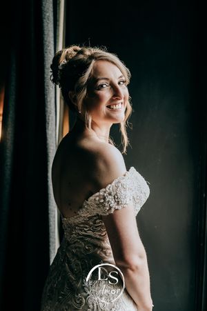 Natural window light illuminates someone wearing an elegant lace wedding dress in a moody dark setting.