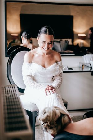 A happy bride in an off-shoulder lace wedding dress sits in a salon chair during wedding preparations.