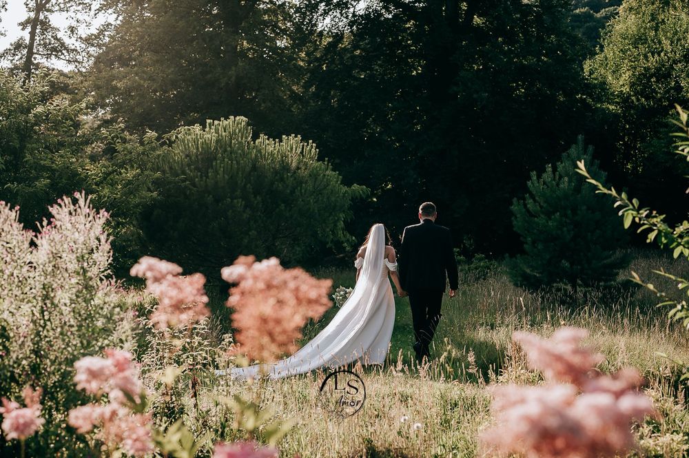 A white wedding dress hangs from a tree in a romantic outdoor garden setting with pink flowers in the foreground.