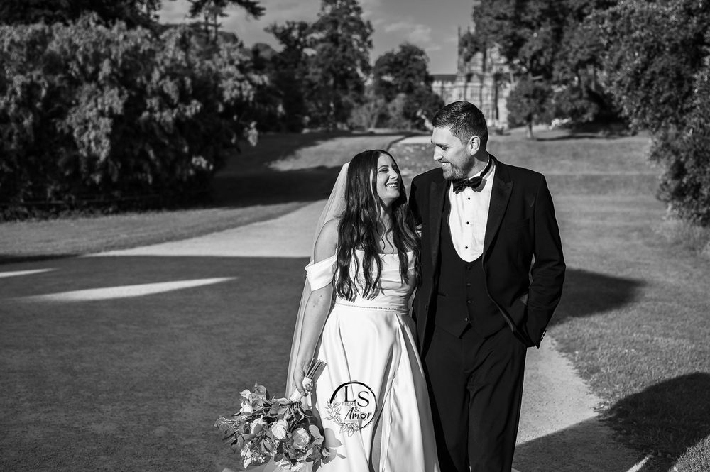 A couple in formal wedding attire walks together on a path through a scenic outdoor garden setting.