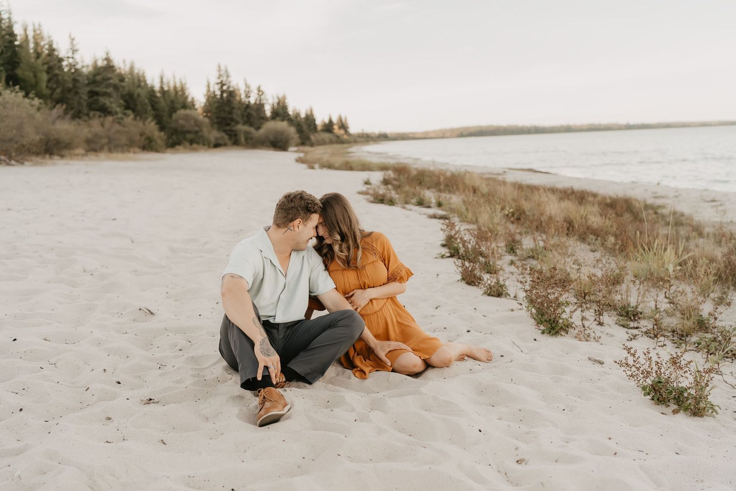 Couple sits together on sandy beach near evergreen forest during sunset.