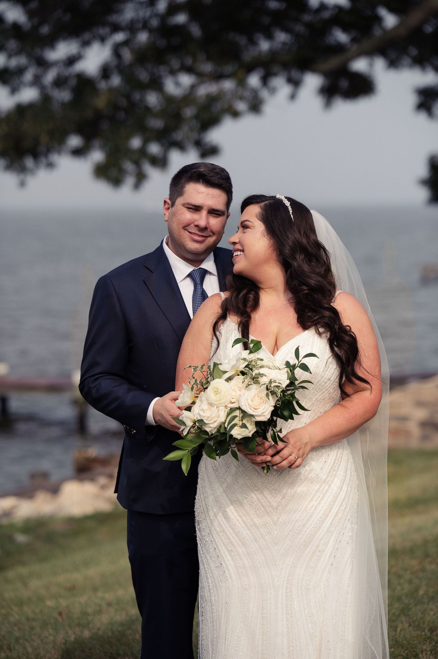 Newlyweds pose with white floral bouquet against waterfront backdrop.