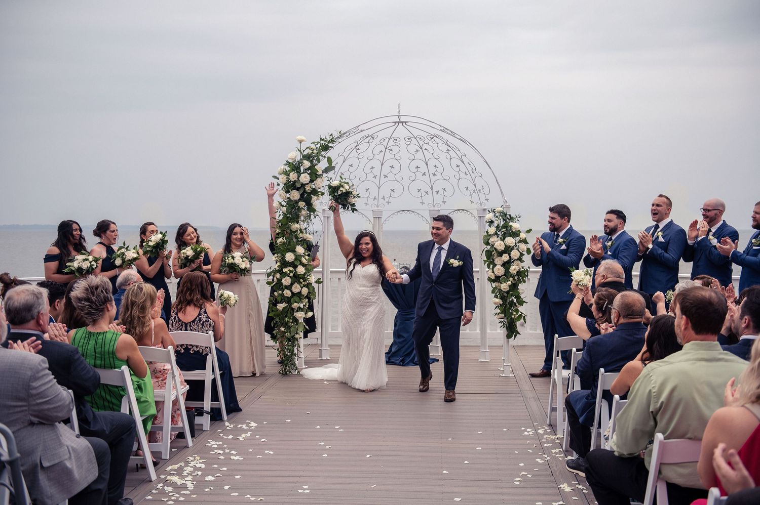 Wedding couple exits ceremony under white metal gazebo as guests cheer.