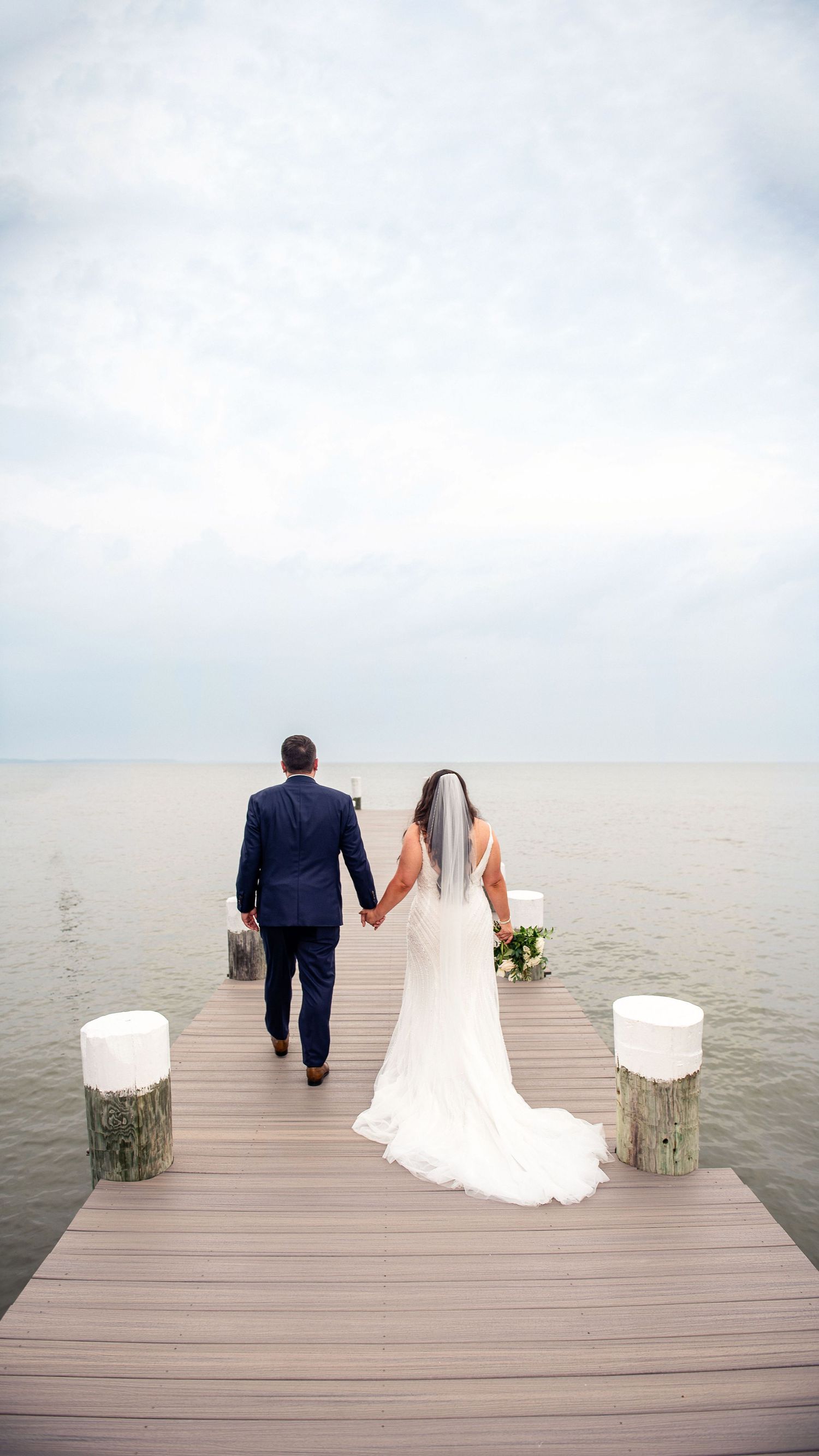 Newlyweds walk hand in hand on wooden pier with white pillar candles.