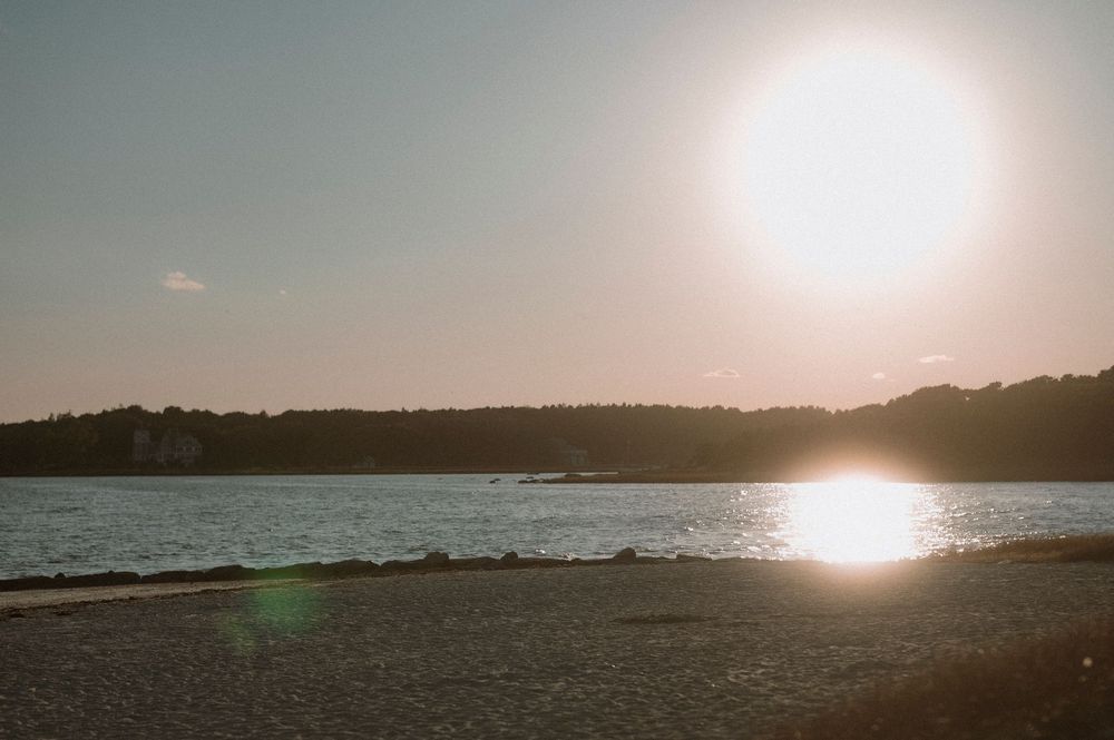 Sun glints off tranquil water at dusk, casting reflections on a peaceful beach with distant tree-lined shore.