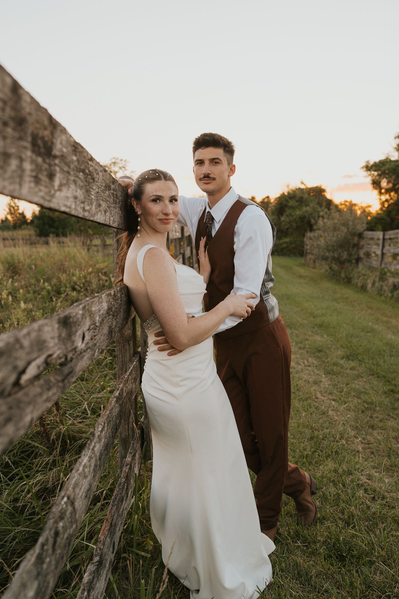 Romantic sunset photoshoot of a married couple embracing near a rustic fence in a grassy field at the Historic Blum Barn in Manchester MI