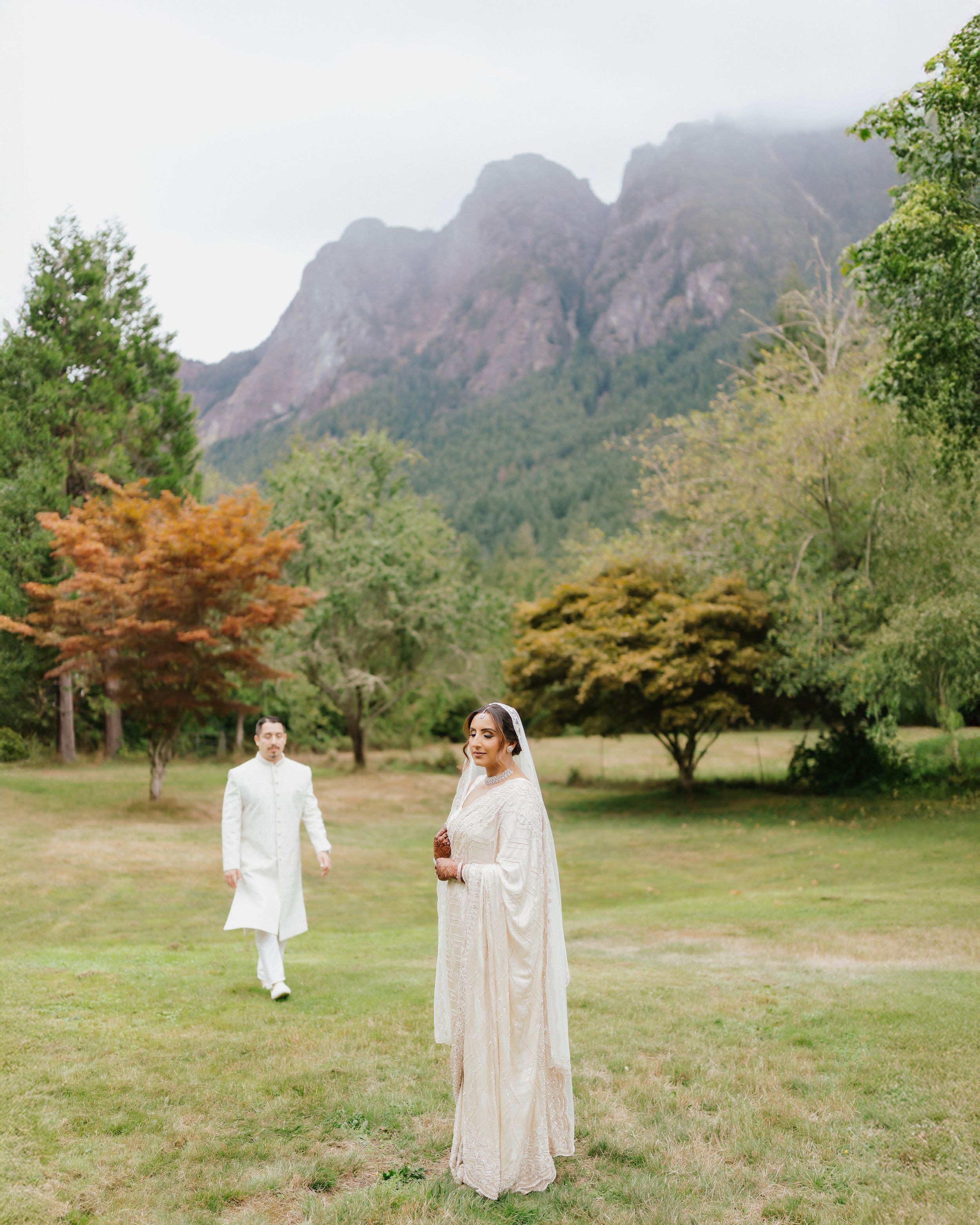 Couple in white outfits walking through a scenic mountain meadow with autumn trees and rugged peaks in the background.