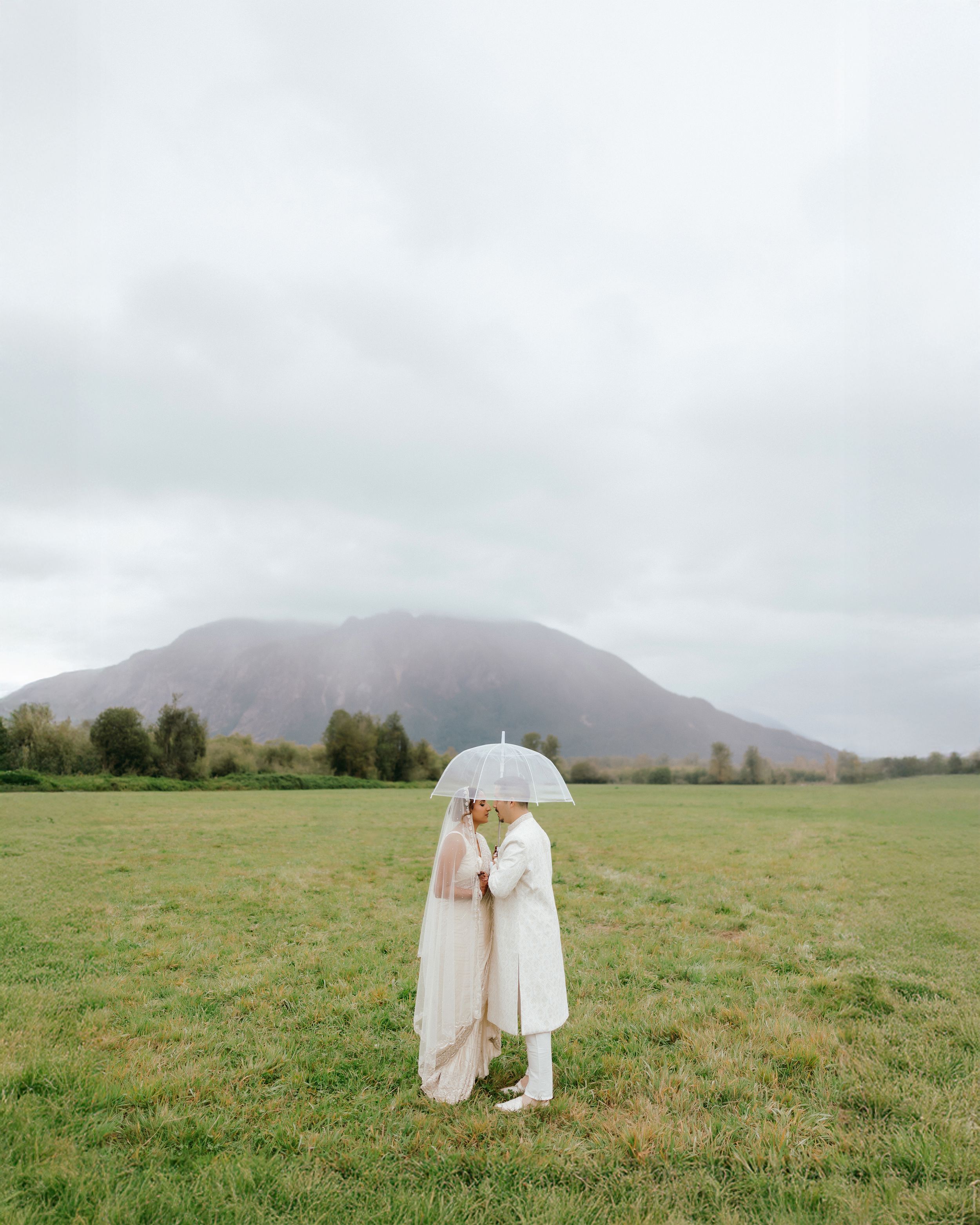 Two people share an umbrella while standing in a misty field with a mountain backdrop on an overcast day.