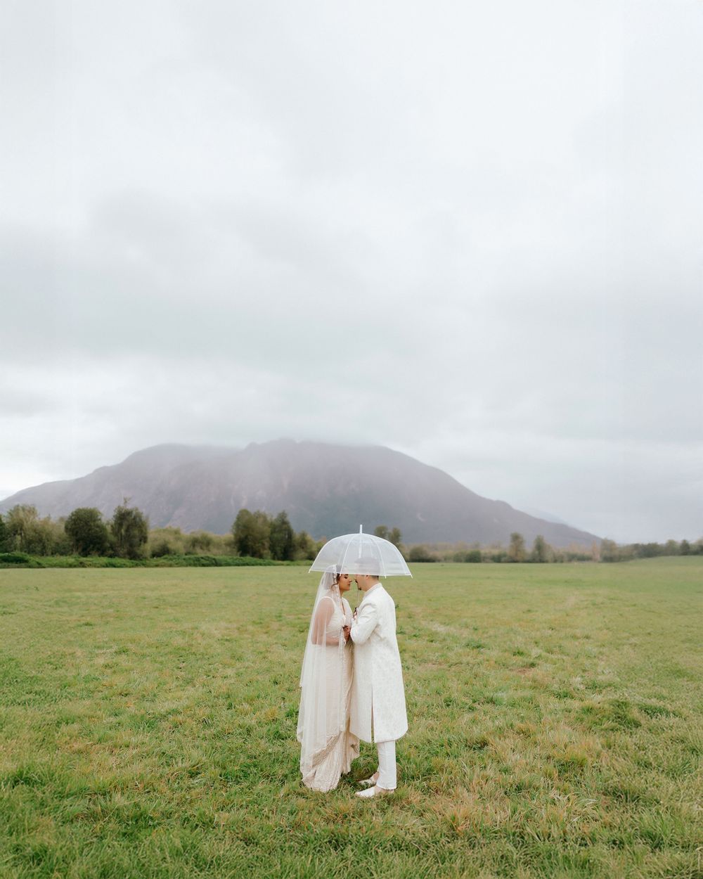Two people share an umbrella while standing in a misty field with a mountain backdrop on an overcast day.