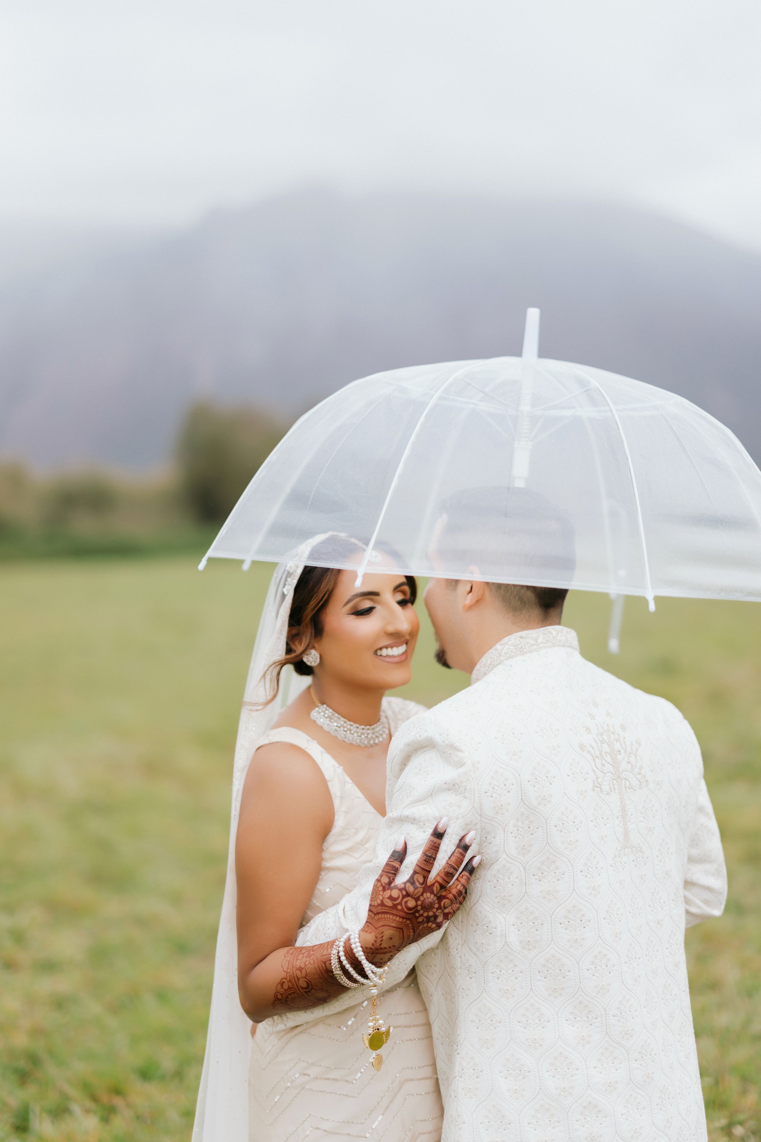 A romantic wedding couple shares an intimate moment under a clear umbrella in a misty field with mountains in the background.