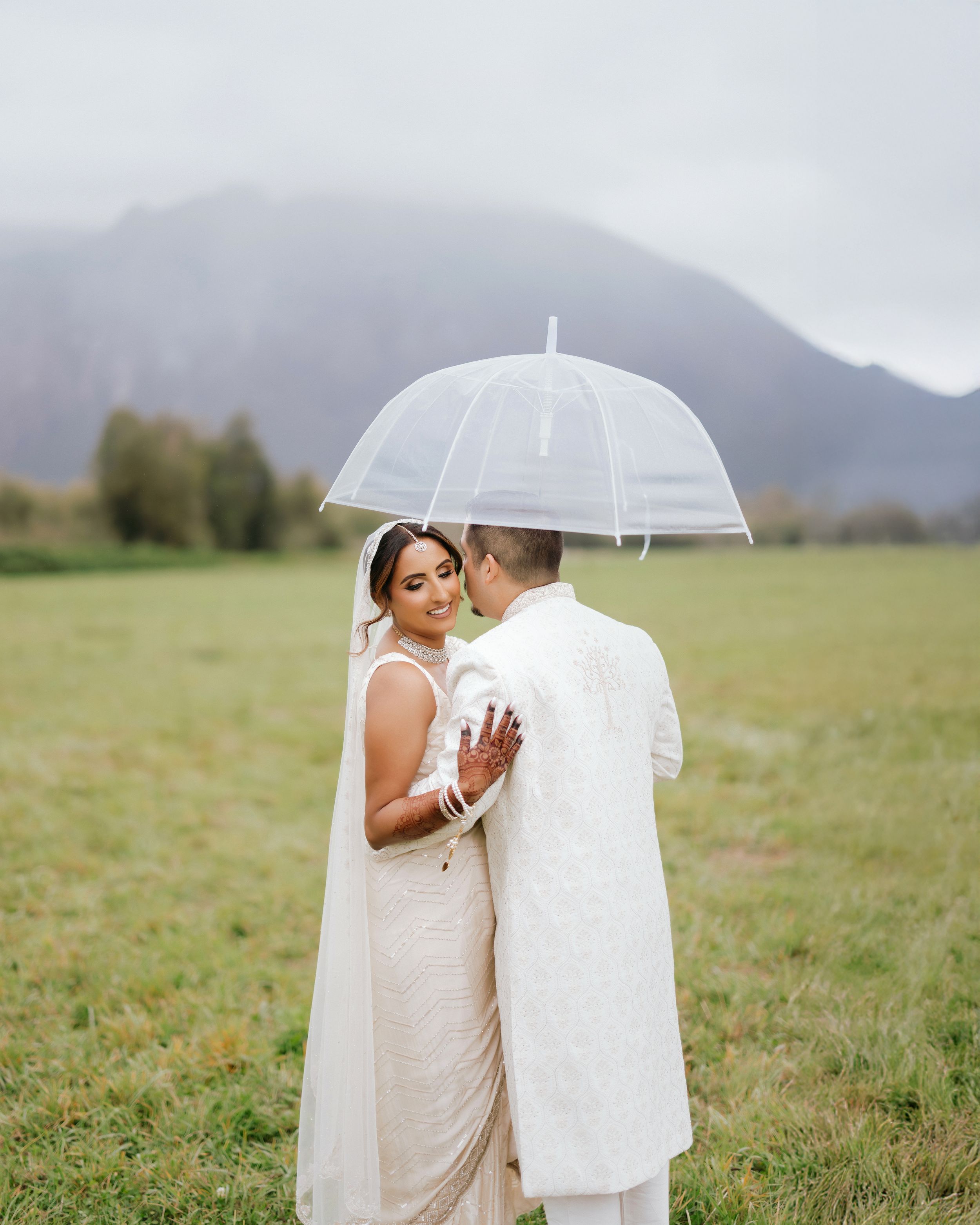 A couple shares an intimate moment under a white umbrella in a misty mountain field on their wedding day.
