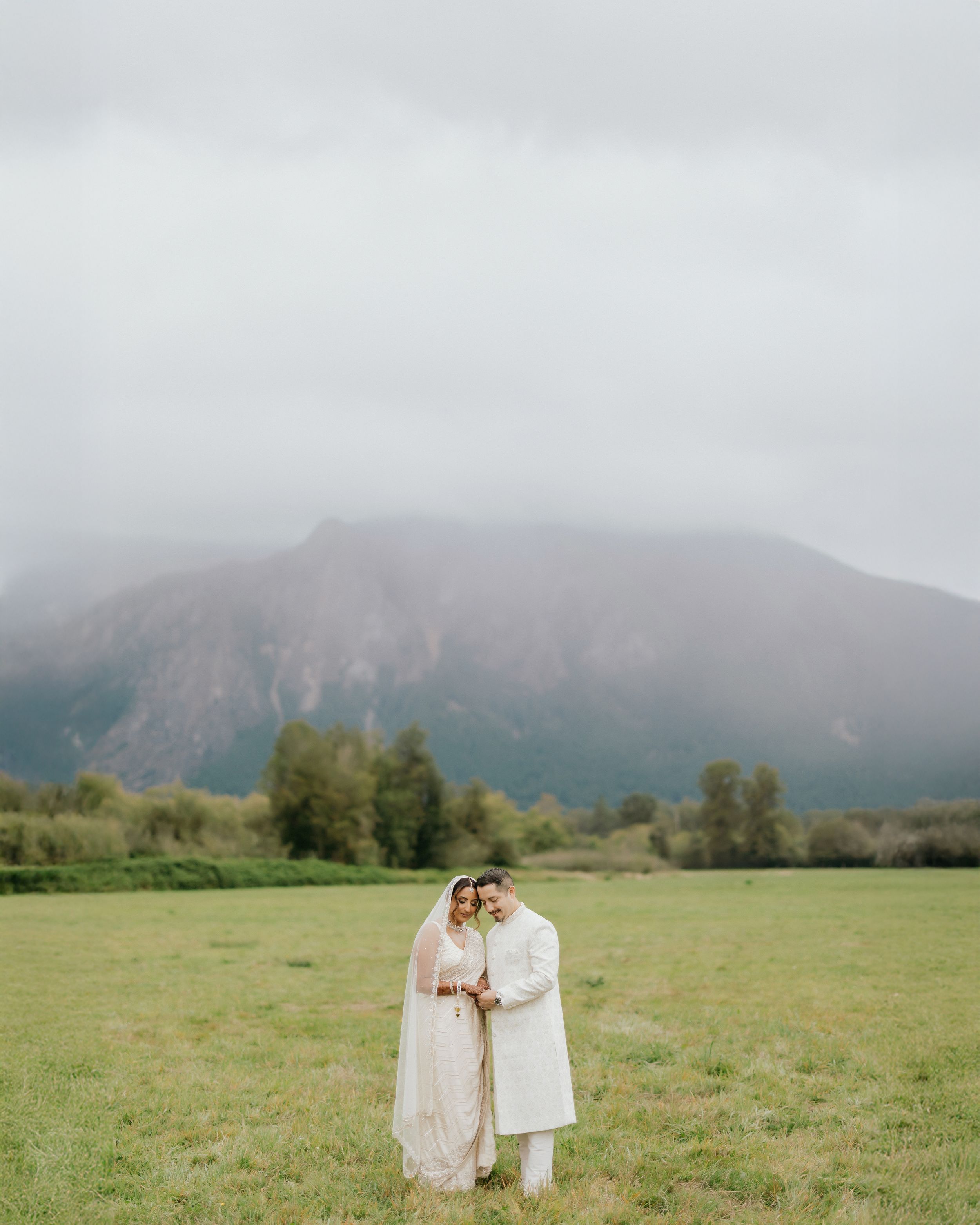 A couple embraces in a serene meadow with misty mountains and evergreen trees in the background.