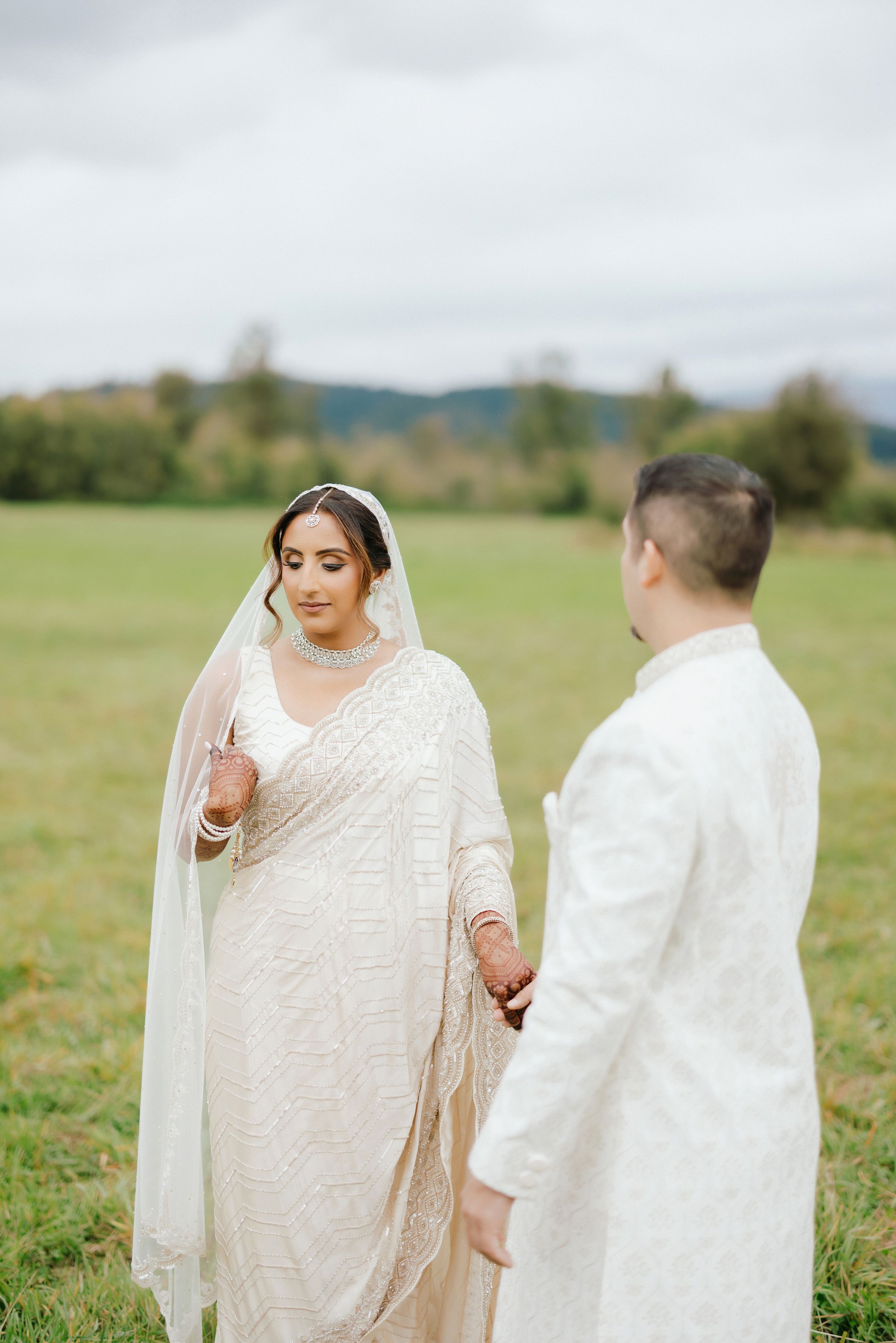 A couple in white attire stands together in a sunny meadow during a romantic outdoor photo session.