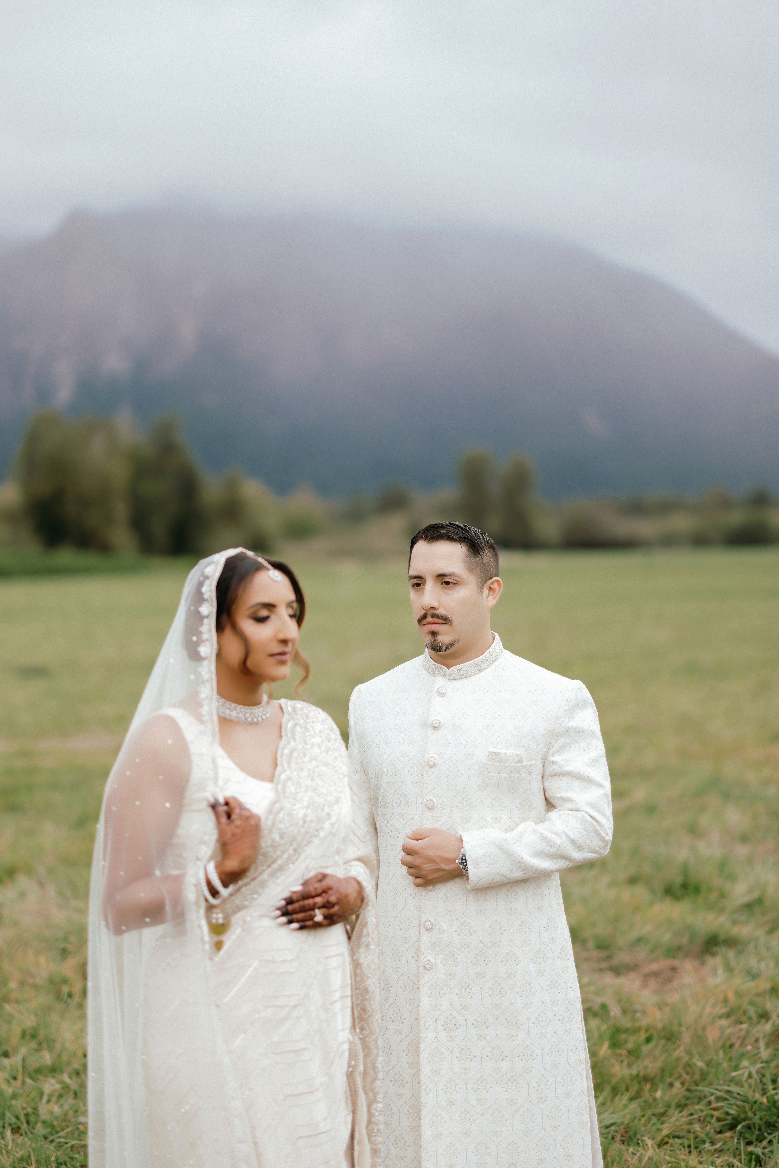 A couple in elegant white wedding attire poses together in a scenic mountain meadow at sunset.