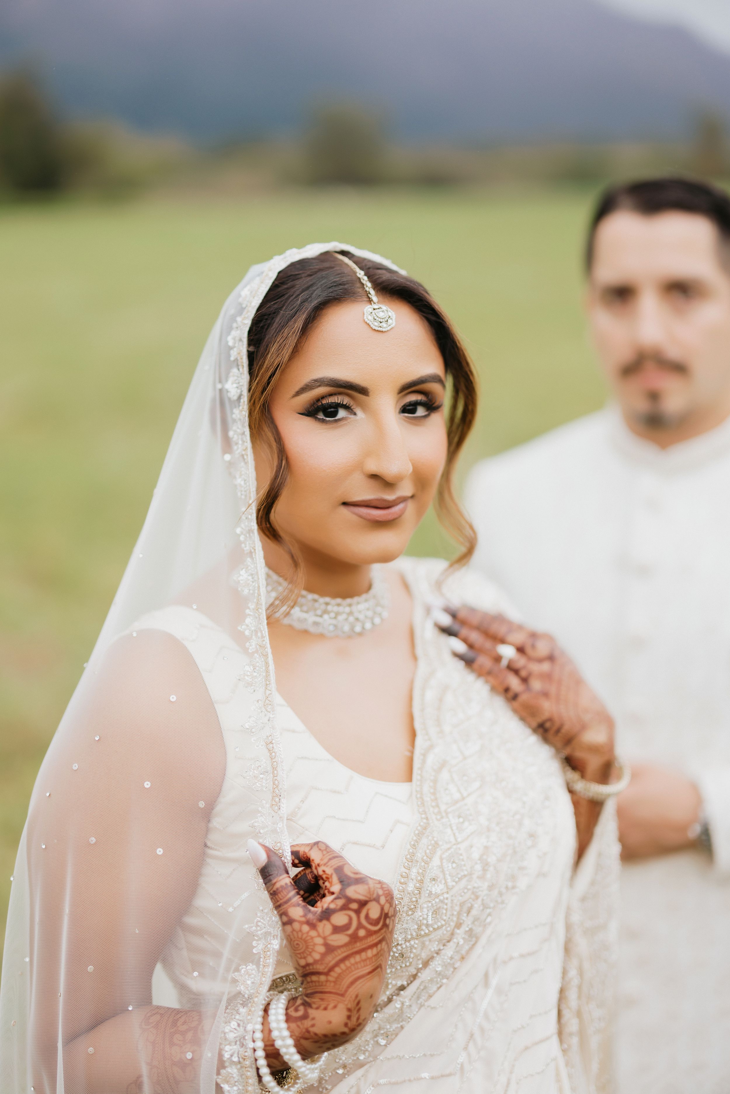 Beautiful bride in traditional South Asian wedding attire with henna and jewelry poses outdoors in natural setting.