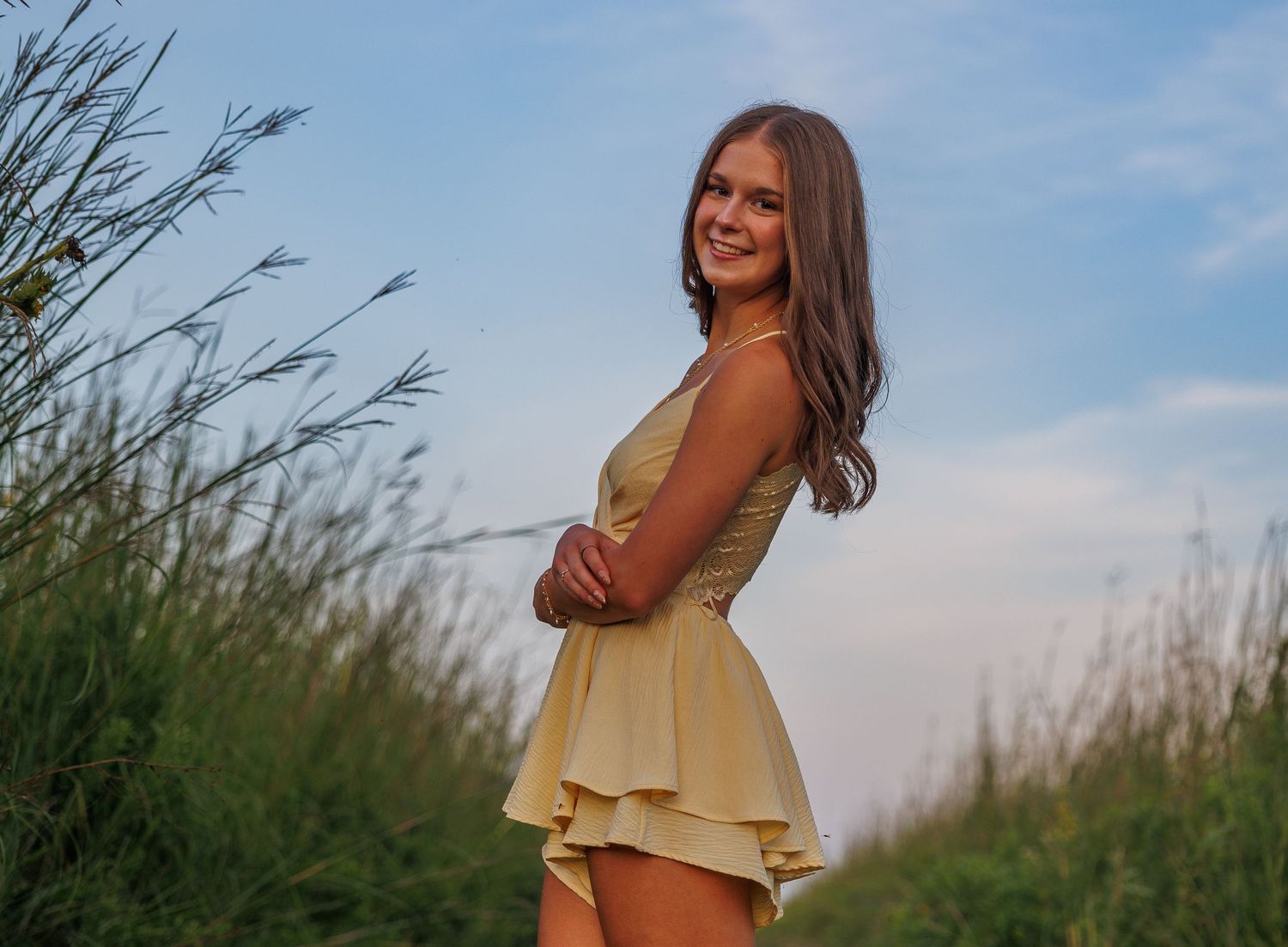 A person wearing a yellow sundress stands in a field of tall grass at sunset against a blue sky.