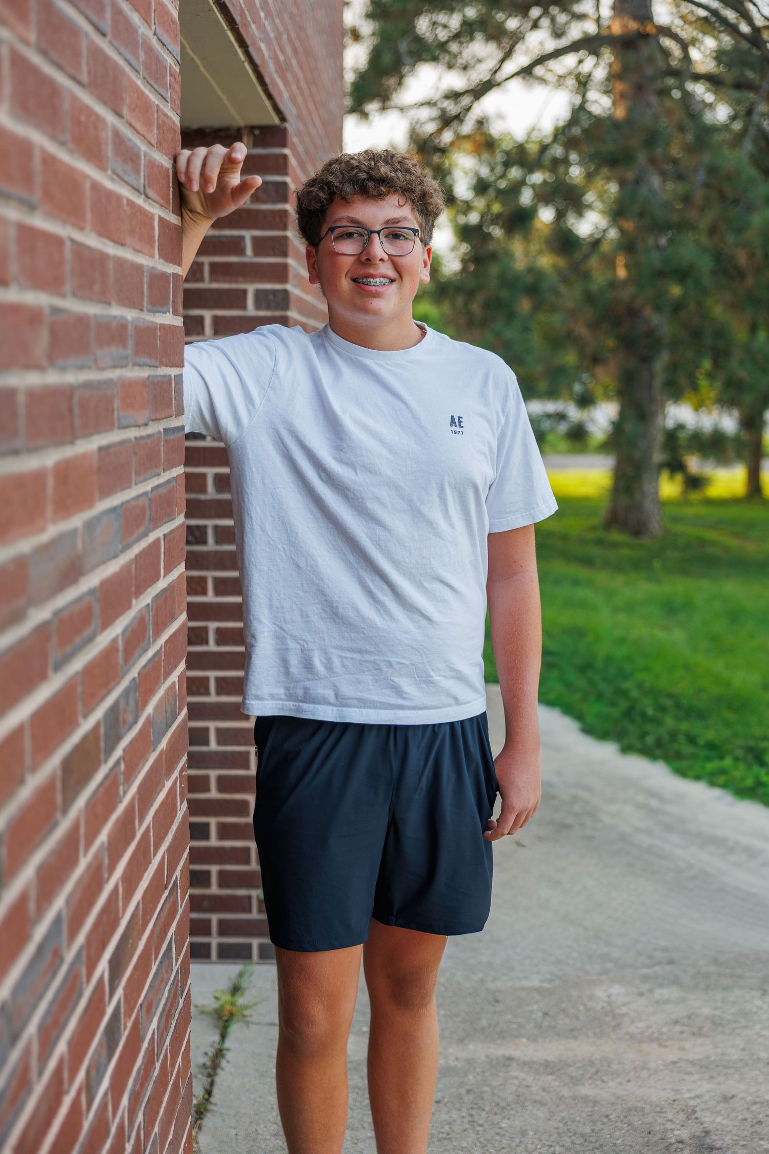A teen in glasses wearing a white t-shirt and navy shorts stands smiling next to a brick building on a sunny day.