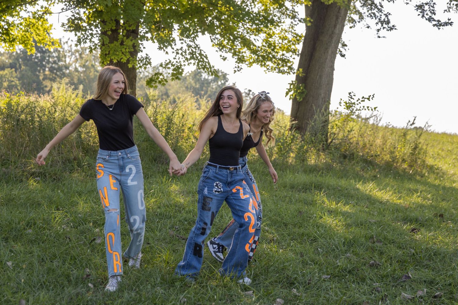Three friends stand together enjoying a sunny day in a grassy field near a large tree.