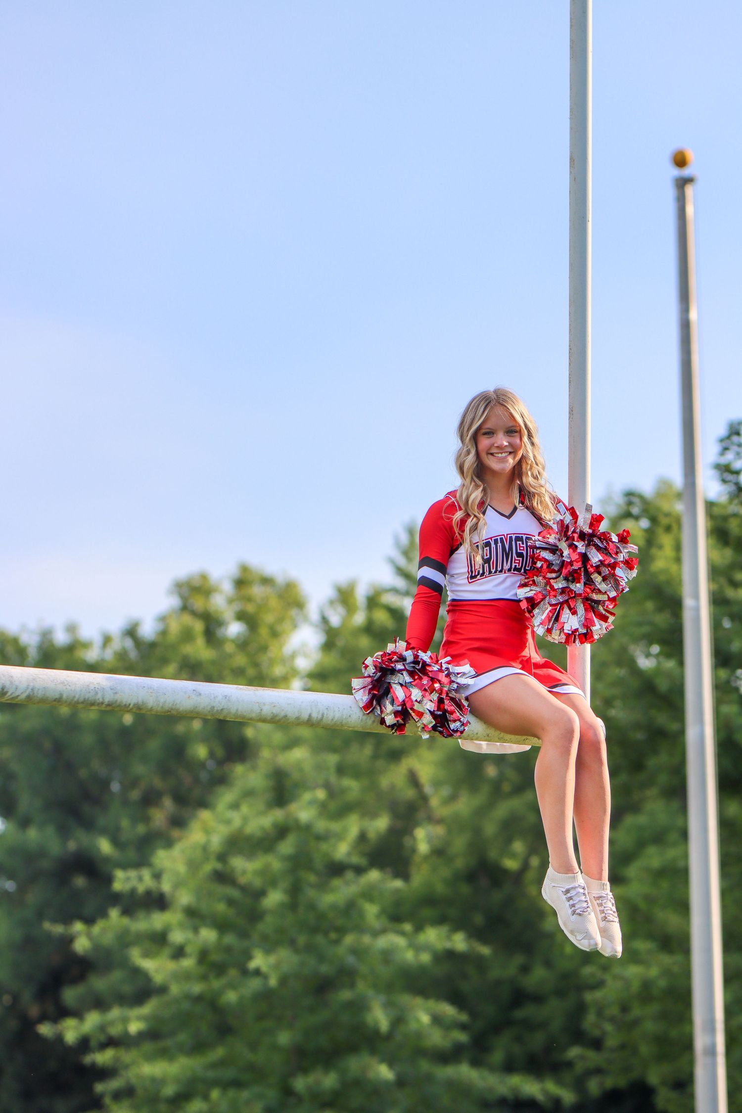 Cheerleader in red uniform sitting on goalpost with pom poms against blue sky.
