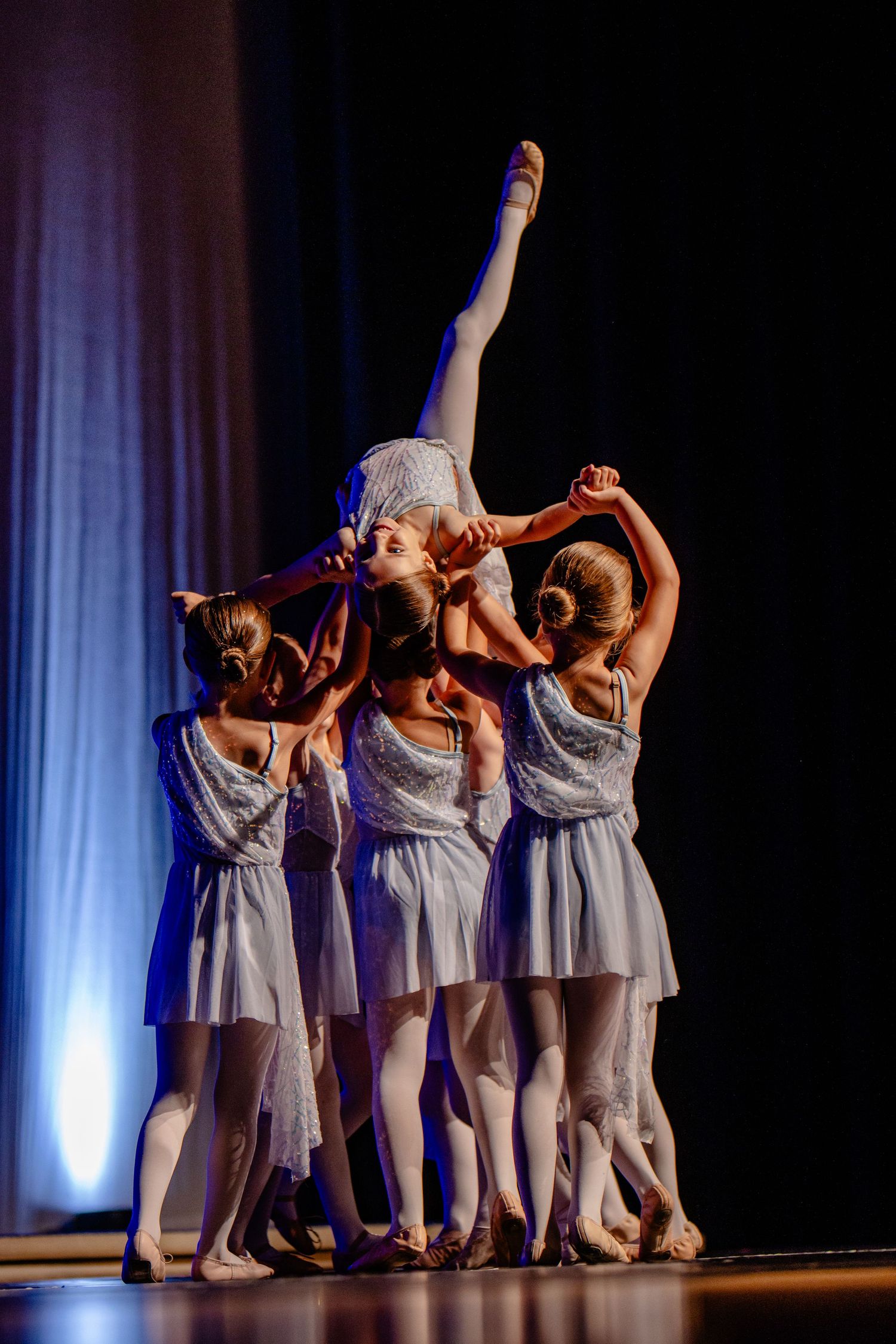 Modern dancers in white dresses performing lifting routine on dark stage.