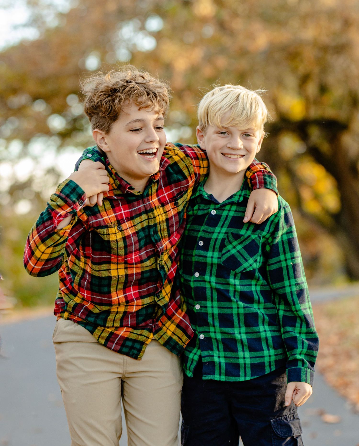 Two children in plaid shirts smile and embrace while posing outdoors during autumn.