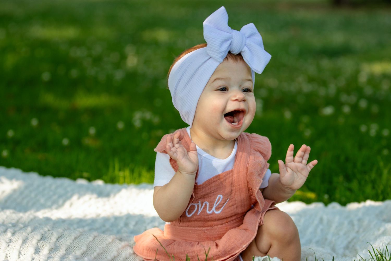 Happy baby in peach romper and grey bow headband laughing on white blanket.