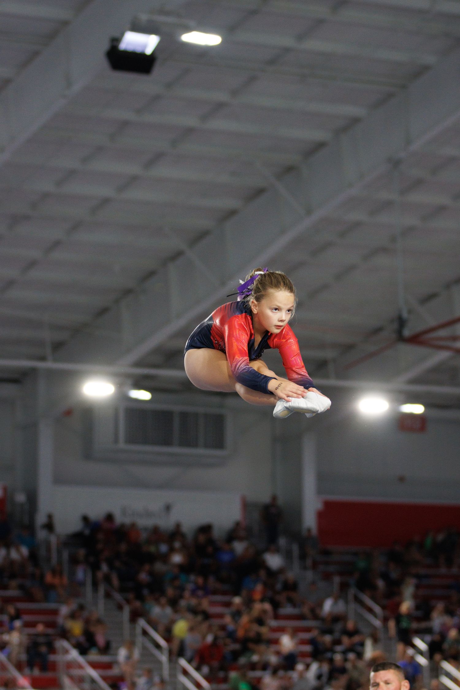 Gymnast in red leotard performs mid-air jump during competition in indoor gymnasium with high ceilings and crowd below.