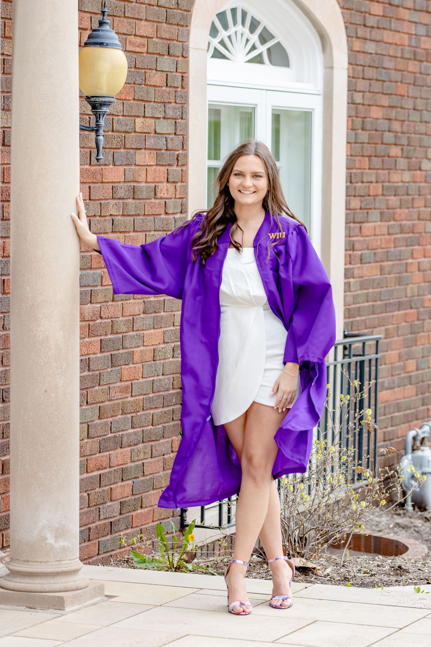 Graduate in purple graduation gown poses against brick building with arched doorway.