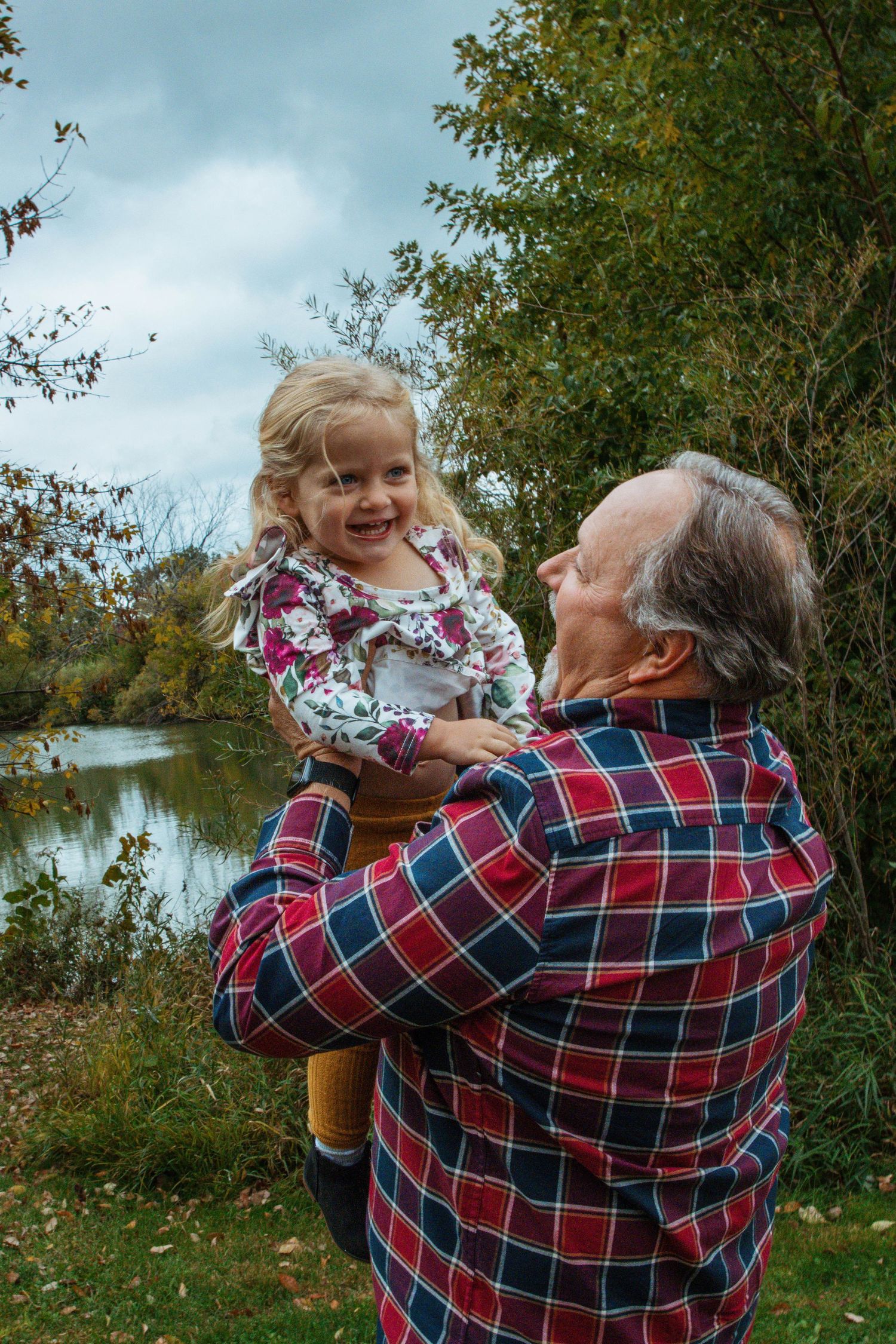 Joyful moment shared between a young child and older adult in plaid shirt by autumn lake.