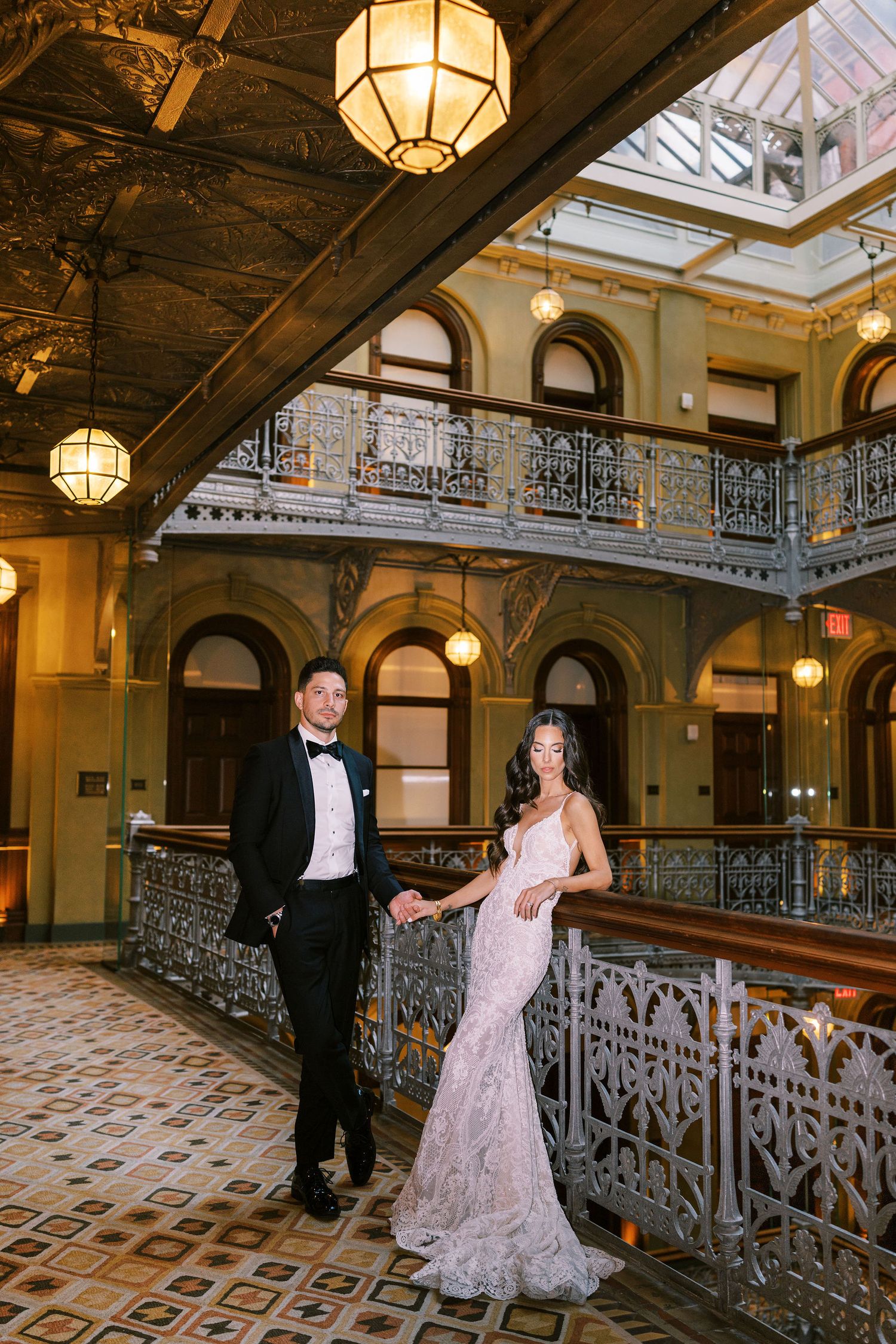 Elegant wedding couple holding hands on ornate iron balcony inside grand historic building with chandeliers.