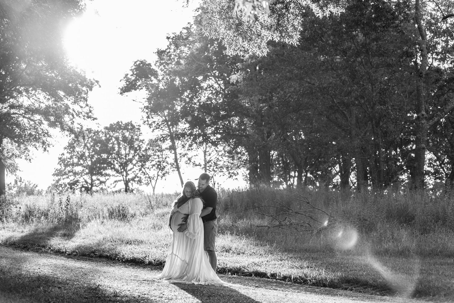 A couple embracing in a field at sunset with pine trees silhouetted against the sky.