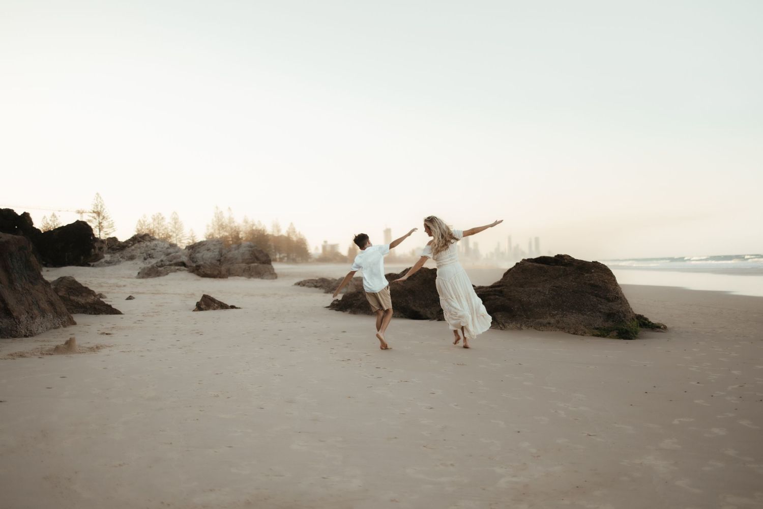Two figures in white dresses dancing together on a misty beach at sunset.