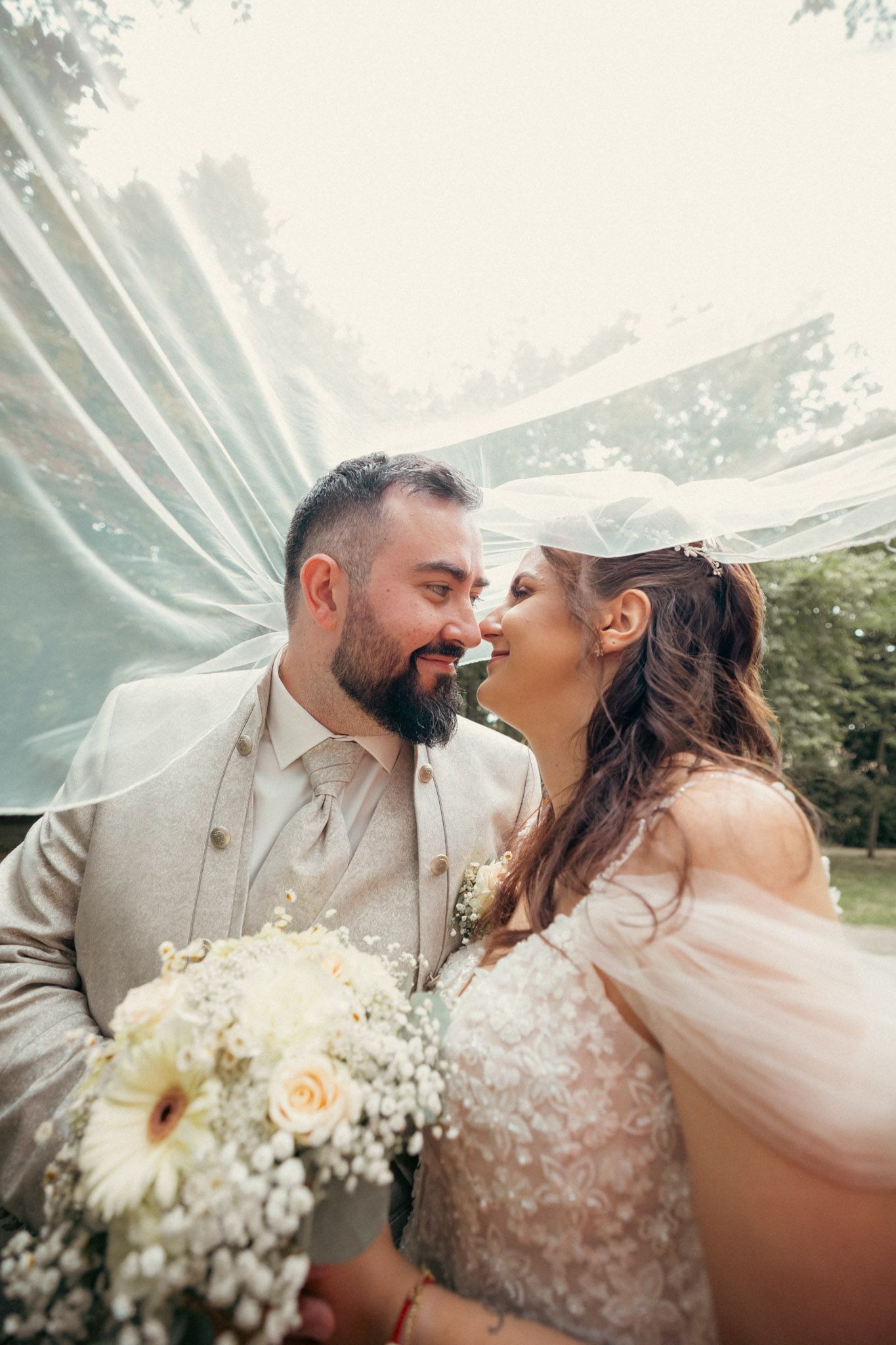 Bride and groom share a romantic moment under a flowing veil, holding a white floral bouquet outdoors.