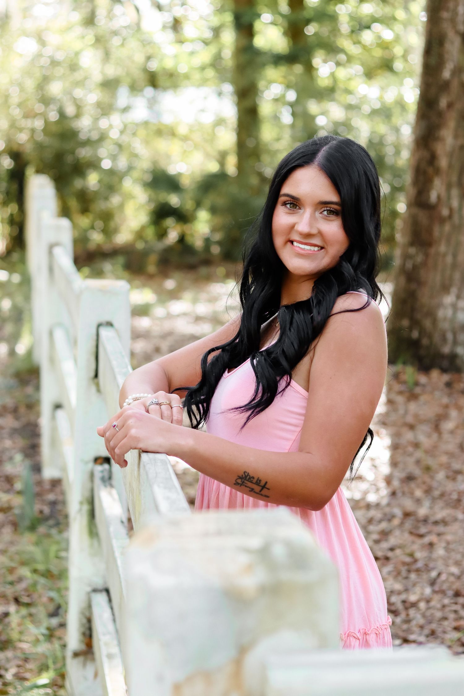 Woman in pink dress poses against rustic white fence in outdoor woodland setting.