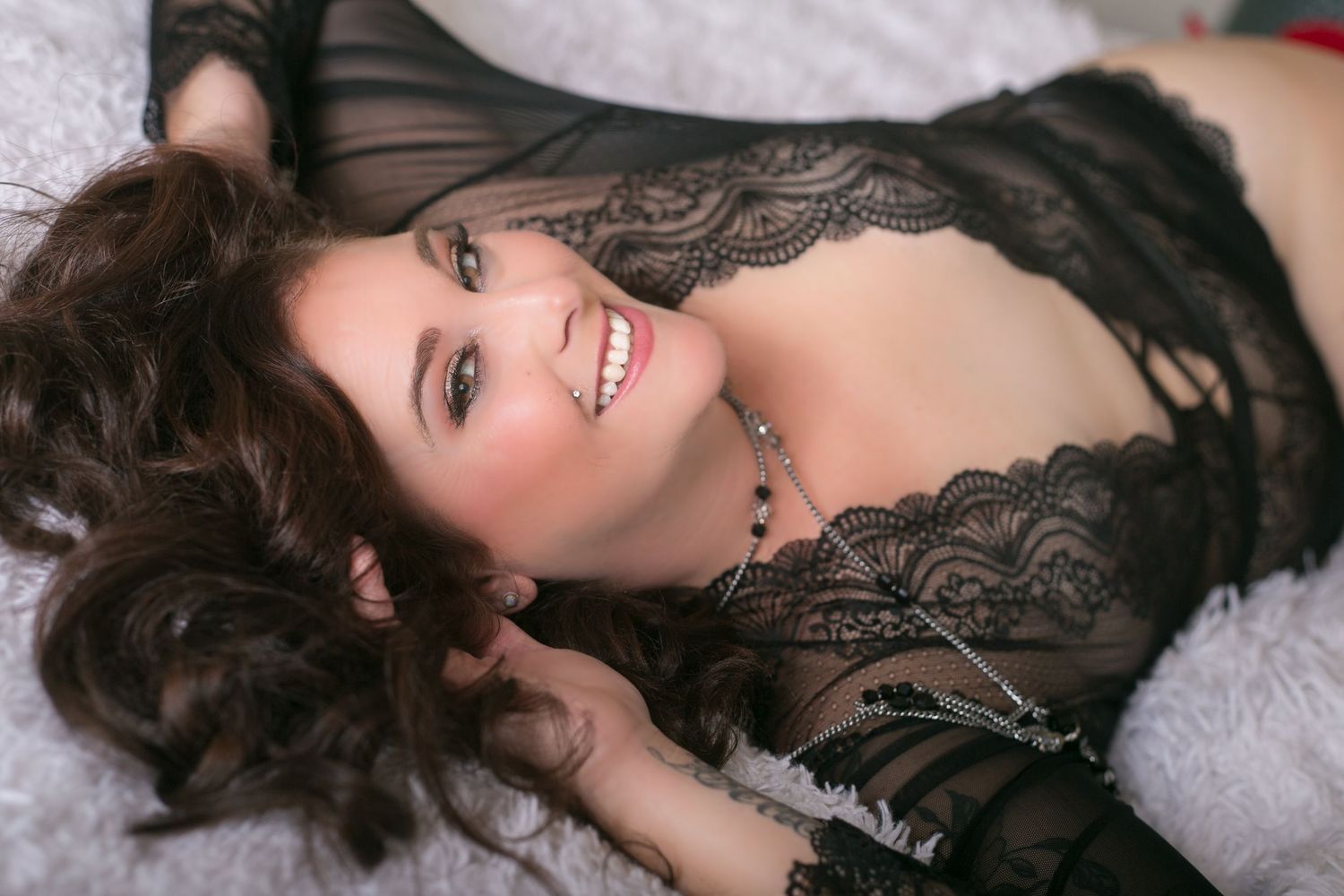 Smiling brunette woman in black lace lingerie lying on white bedding looking at camera.