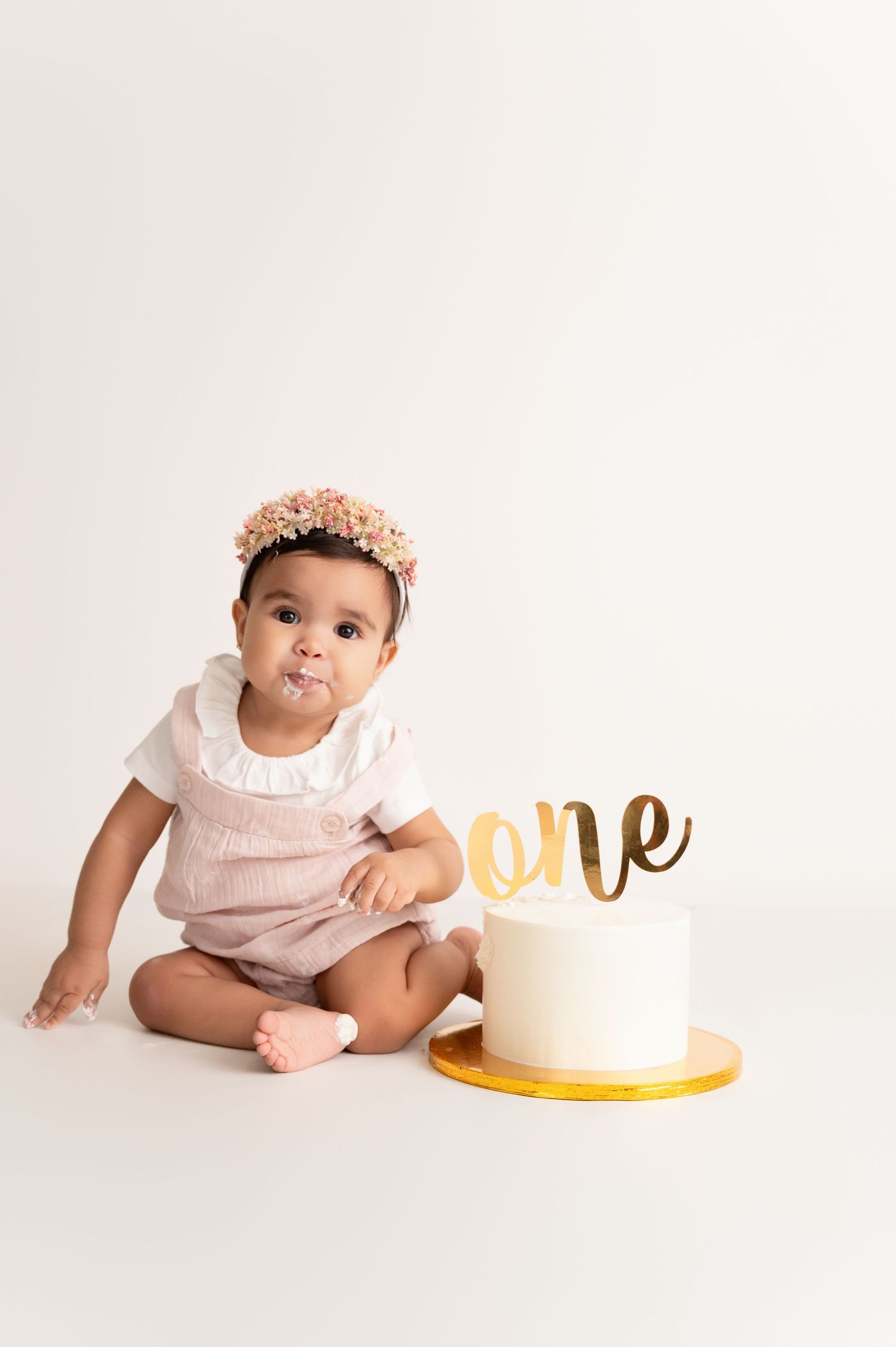 First birthday cake smash session with baby in pink dress next to gold 'one' cake topper.