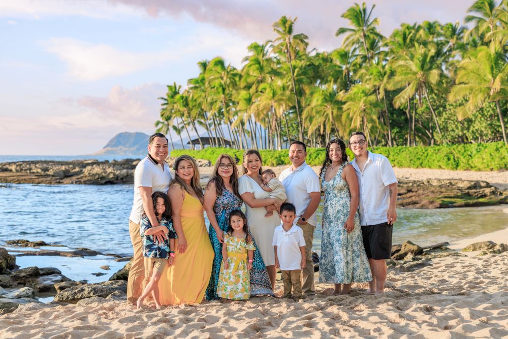 Family portrait on a Hawaiian beach with palm trees, posing together at sunset near rocky shoreline.