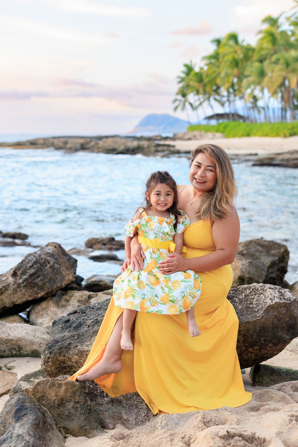 Mother and daughter in matching yellow dresses sitting on rocks at a tropical beach with palm trees at sunset.