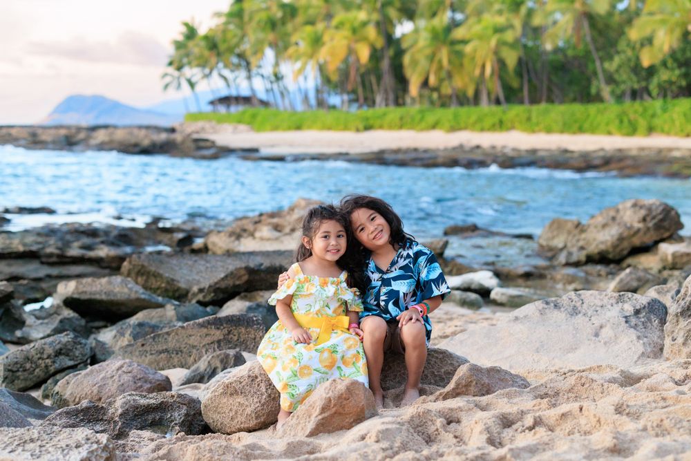 Two children sitting on rocks at a tropical beach with palm trees and ocean in Hawaii.