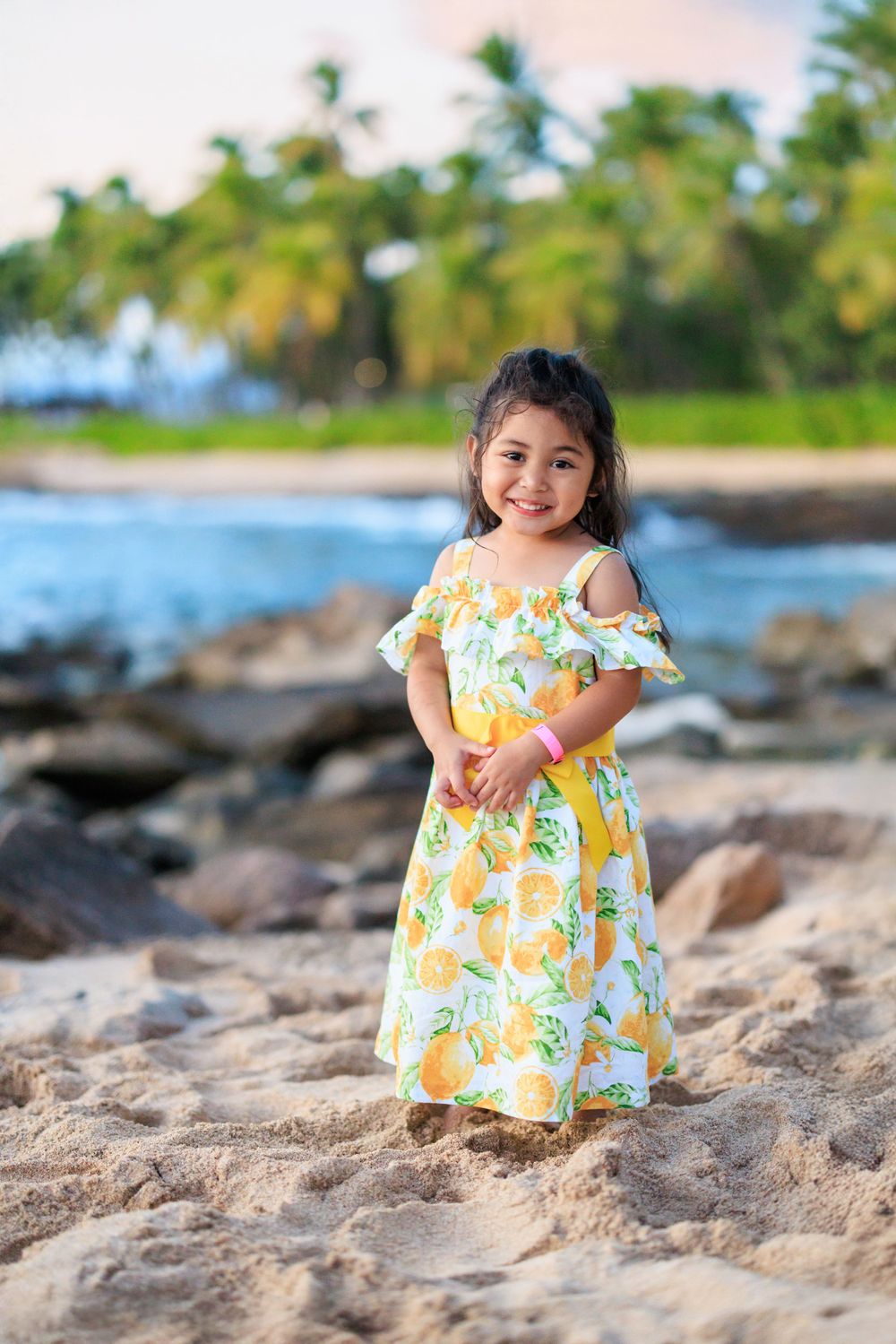 Young girl in floral yellow dress smiling on sandy beach with rocks and palm trees in background.