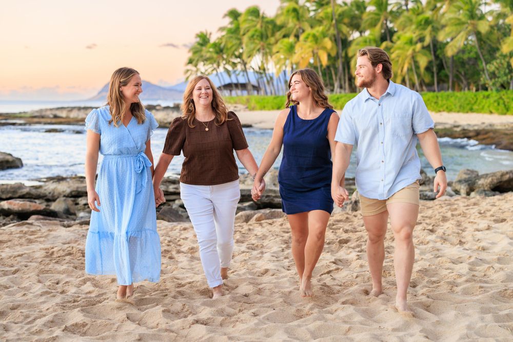 Four people walk hand-in-hand on a sandy Hawaiian beach at sunset with palm trees and ocean in the background.