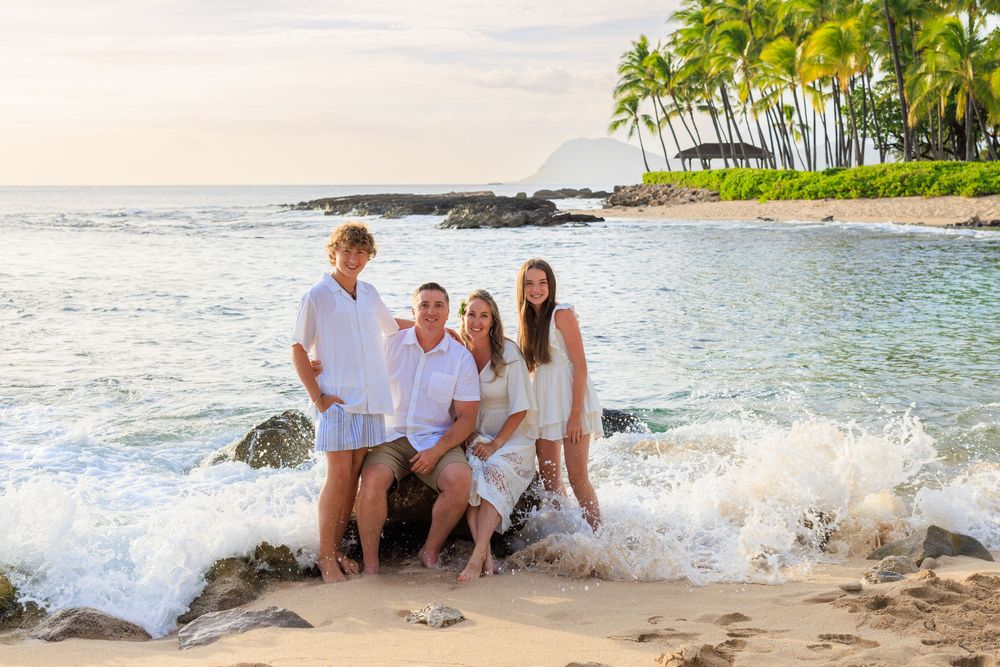 Family of four smiling together on a sandy Hawaiian beach with ocean waves and palm trees in the background.