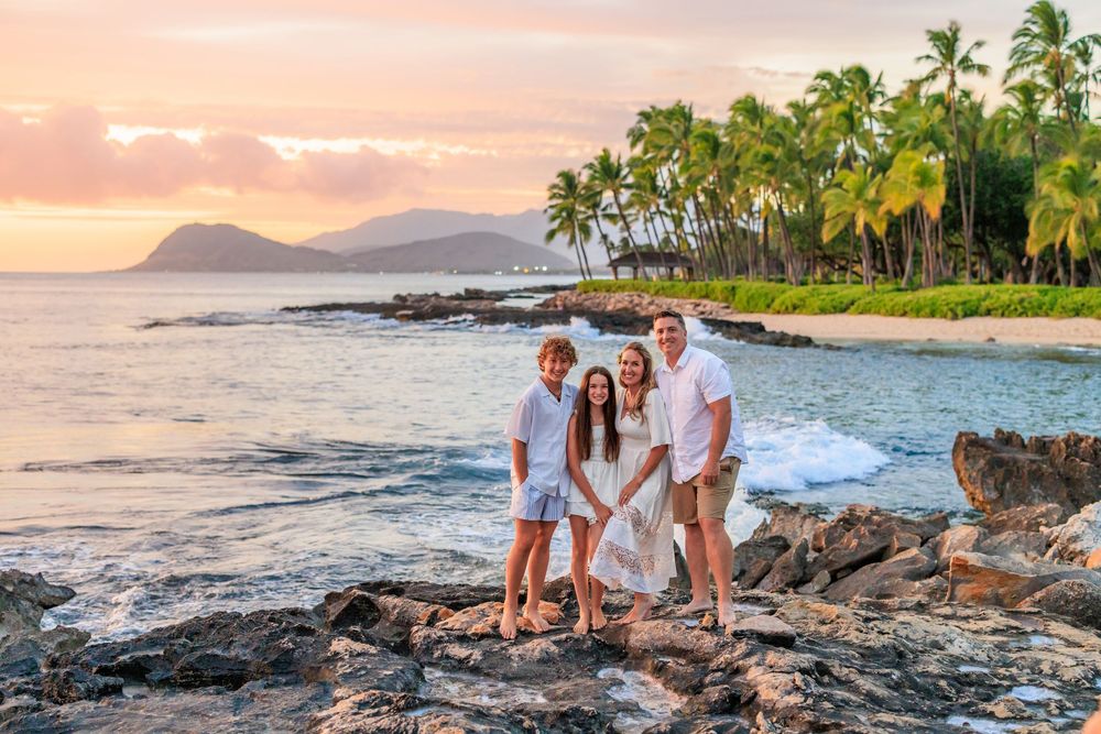 Family of four posing on rocky Hawaiian beach at sunset with palm trees and ocean backdrop.
