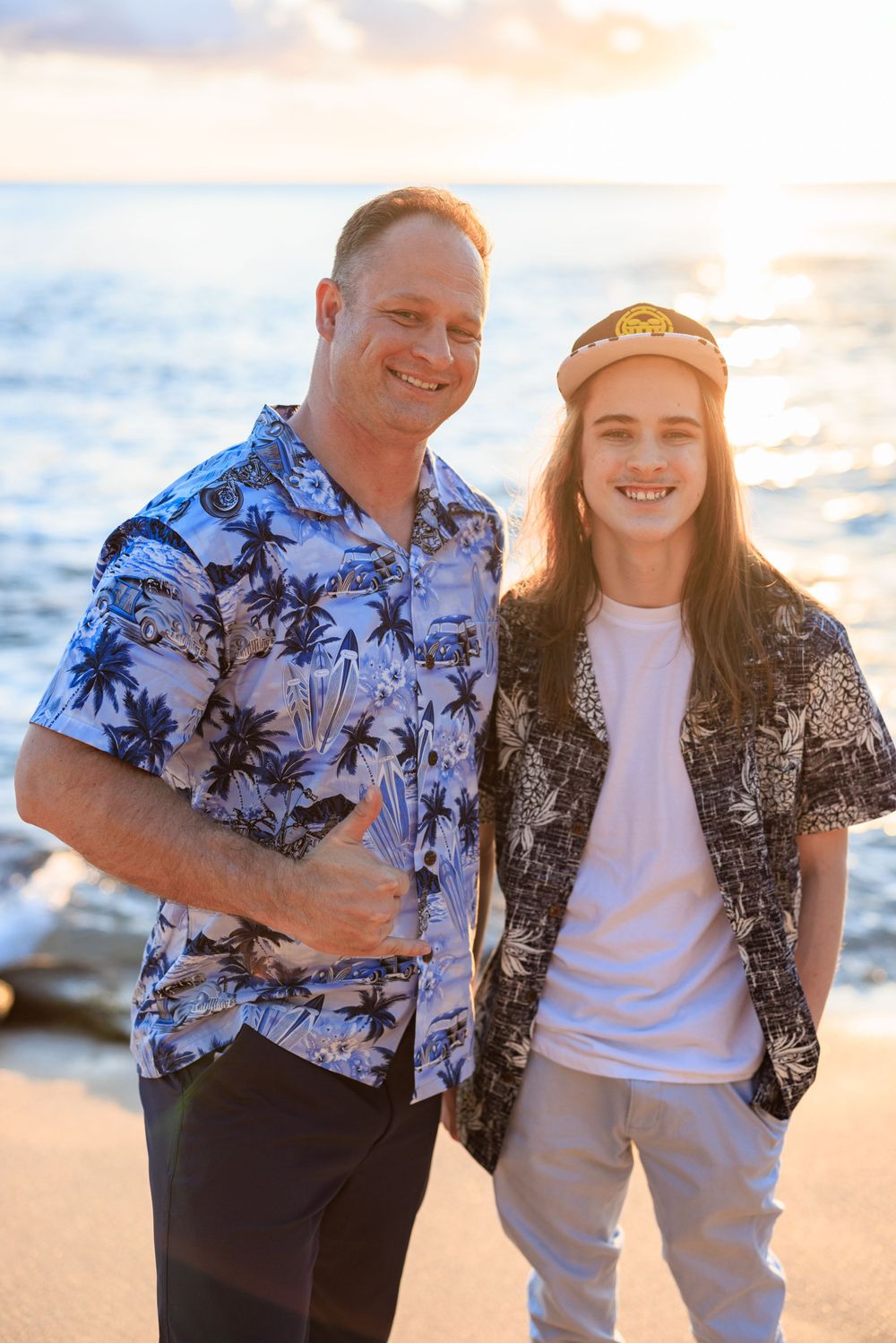 Two smiling males in Hawaiian shirts pose together on a sunny beach at golden hour.