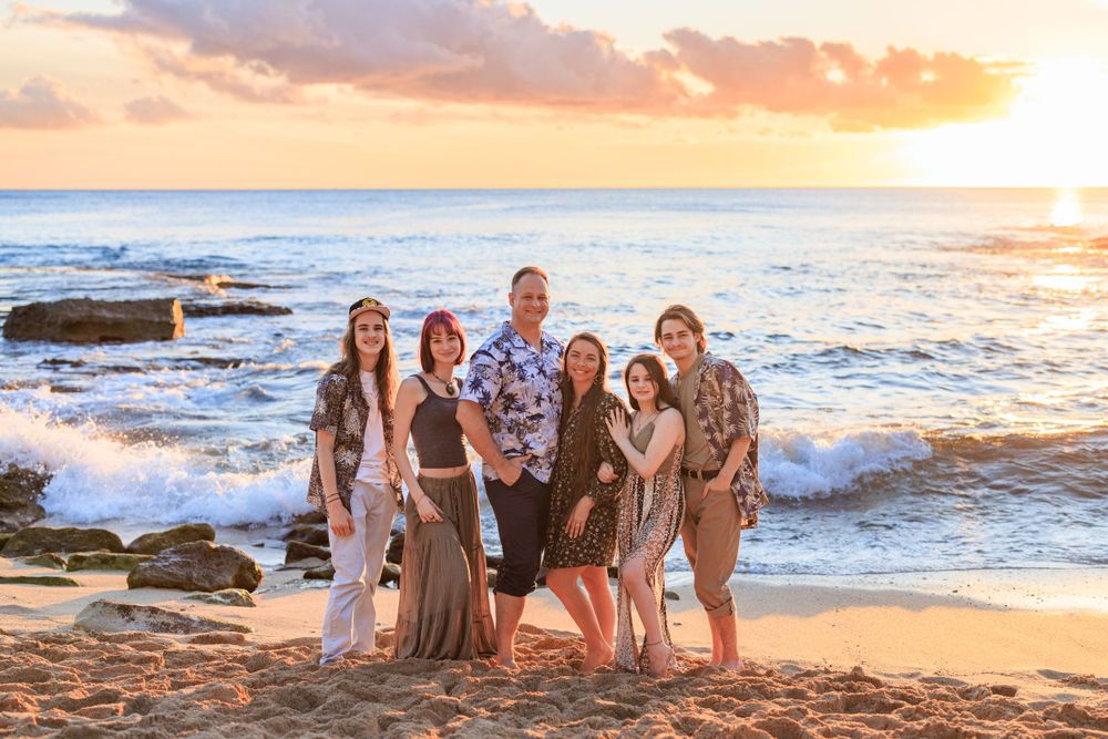 Family of six poses on sandy beach at golden sunset with ocean waves, rocks, and colorful sky backdrop.