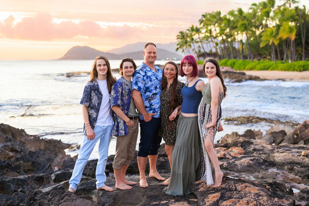 Family of six posing on rocky Hawaiian beach at sunset with palm trees and ocean in background.