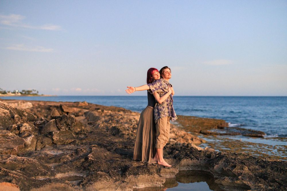 Couple embracing on rocky shoreline at sunset with blue ocean waves and clear sky in background.