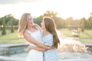 Two blonde women in floral dresses embrace and smile near a fountain at golden hour.
