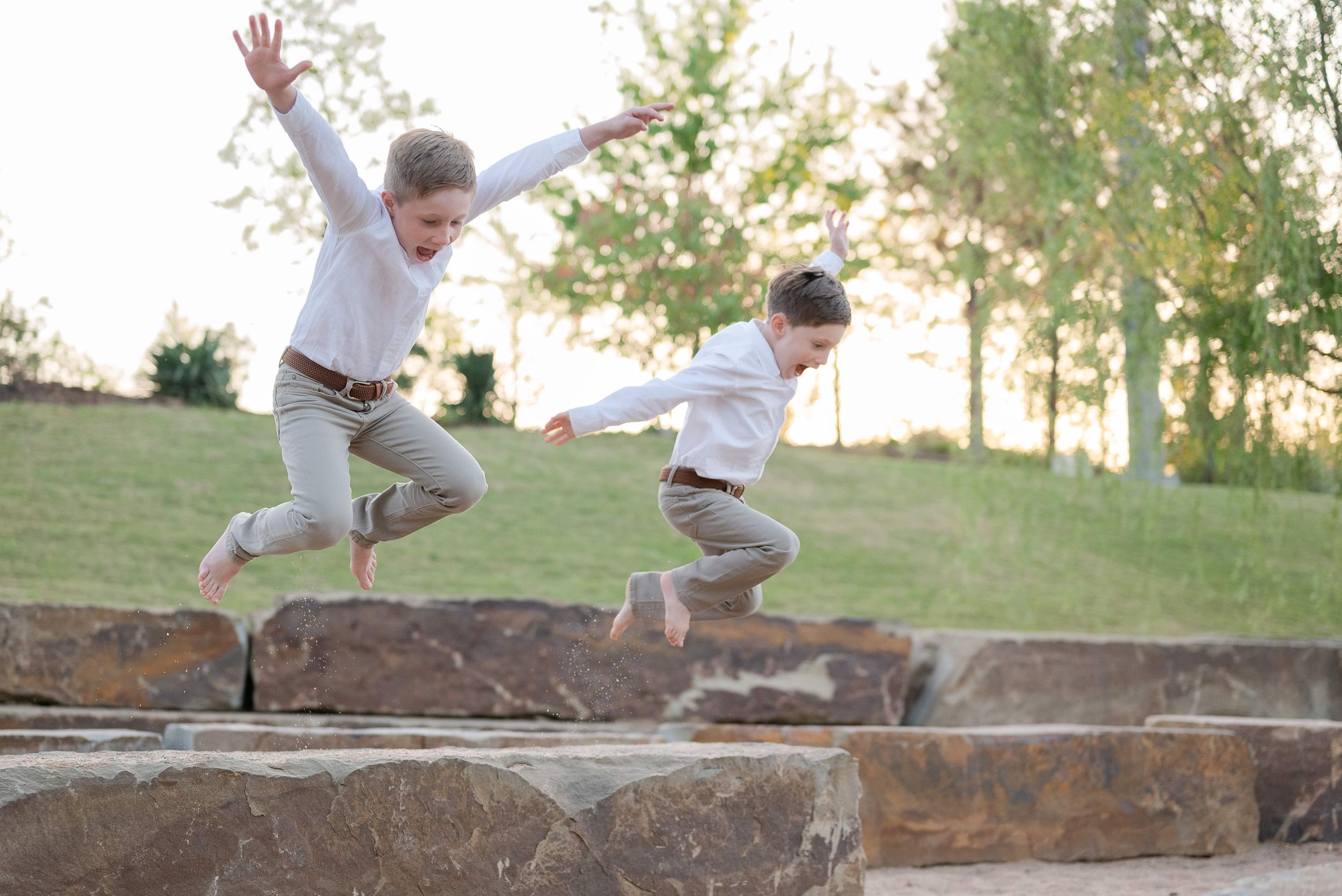 Two children joyfully jumping off stone steps outdoors, wearing matching white shirts and tan pants in a park setting.